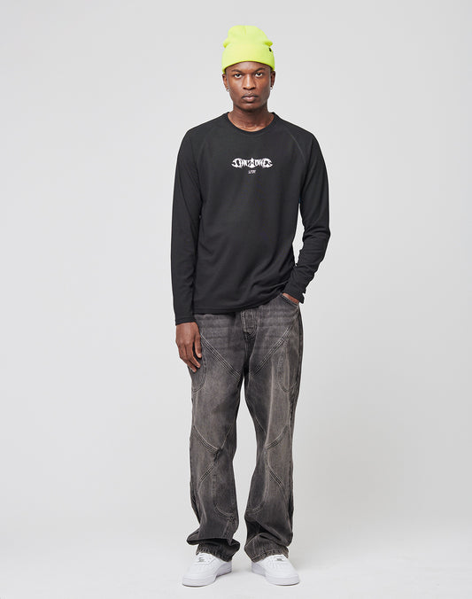A man stands against a white background wearing a neon yellow beanie, the LIVE FAST DIE YOUNG Live Fast Sports Longsleeve in black with a white graphic, faded baggy jeans, and white sneakers. His expression is neutral and his hands are relaxed by his sides.