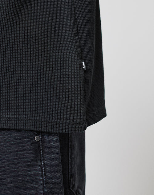 Close-up of the LIVE FAST DIE YOUNG Blank Waffle Longsleeve in black with textured fabric, Ripp-Bündchen, and small side tag, styled with dark denim pants against a plain light backdrop.
