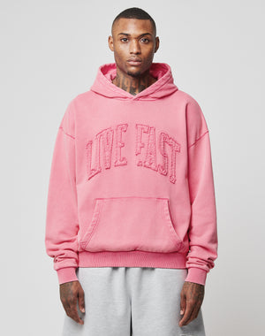 A person wearing a pink Ripped College Hooded sweatshirt by LIVE FAST DIE YOUNG with distressed LIVE FAST lettering and light gray sweatpants, standing against a plain light background.