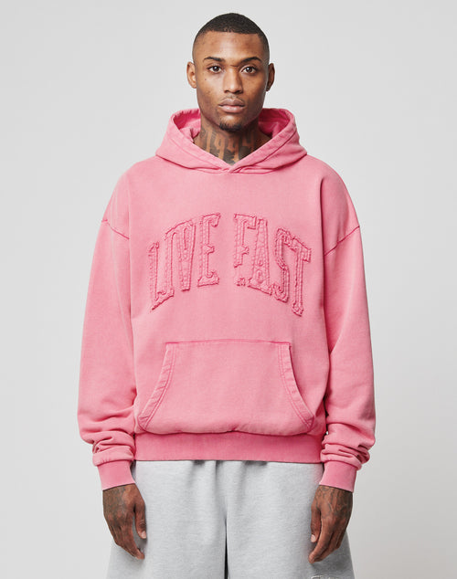 A person wearing a pink Ripped College Hooded sweatshirt by LIVE FAST DIE YOUNG with distressed LIVE FAST lettering and light gray sweatpants, standing against a plain light background.