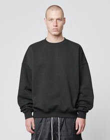 A person with a buzz cut is wearing the LIVE FAST DIE YOUNG Basic Blank Sweater, an Oversize Fit black sweatshirt made from soft Baumwolle and Polyester, paired with black shorts, standing against a plain light gray background.