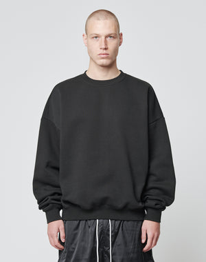 A person with a buzz cut is wearing the LIVE FAST DIE YOUNG Basic Blank Sweater, an Oversize Fit black sweatshirt made from soft Baumwolle and Polyester, paired with black shorts, standing against a plain light gray background.
