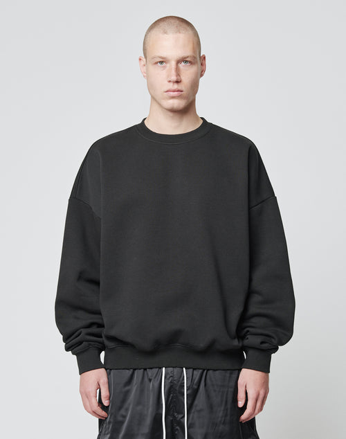 A person with a buzz cut is wearing the LIVE FAST DIE YOUNG Basic Blank Sweater, an Oversize Fit black sweatshirt made from soft Baumwolle and Polyester, paired with black shorts, standing against a plain light gray background.