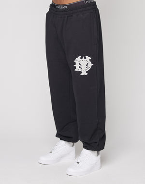 A person wears DY Loose Jogger by LIVE FAST DIE YOUNG—black cotton pants with a white abstract logo on the left thigh, an elastic waistband with "LIVE FAST" text, paired with white sneakers, standing against a plain white background.