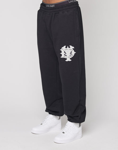 A person wears DY Loose Jogger by LIVE FAST DIE YOUNG—black cotton pants with a white abstract logo on the left thigh, an elastic waistband with "LIVE FAST" text, paired with white sneakers, standing against a plain white background.