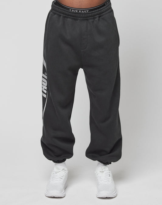 A person wears LIVE FAST DIE YOUNG’s LFDY Sphere Jogger, black with a white left-leg graphic and an Elastikbund waistband reading LIVE FAST, paired with white sneakers against a plain white background.