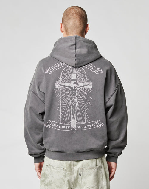 Someone seen from behind wears a dark gray LFDY Cross Hooded Zip by LIVE FAST DIE YOUNG, featuring a large crucifix graphic and LIVE FOR IT OR DIE BY IT on the back with a Distress-Effekt, paired with light-colored pants.