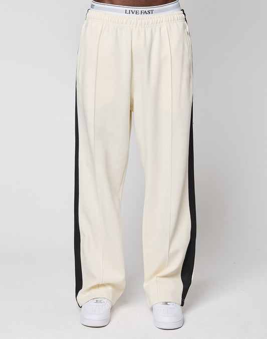 A person wears cream LFDY Striped Jogger by LIVE FAST DIE YOUNG, featuring black side stripes and an elastic waistband with "LIVE FAST." Paired with white sneakers, the waist-down photo is set against a plain background.