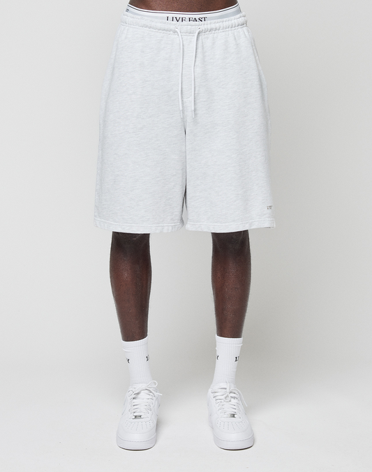 A person wears LIVE FAST DIE YOUNG Basic Shorts in light gray with elastic waistband and "LIVE FAST" logo print, along with white crew socks and sneakers, standing against a plain white background. The upper body is not visible.