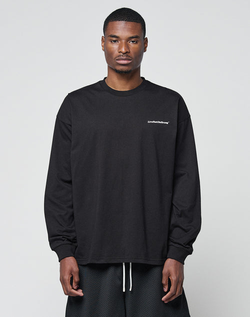 A man faces forward in a LIVE FAST DIE YOUNG Basic Longsleeve—black, loose-fit, with subtle logo text on the left chest—paired with black shorts. The oversize style looks relaxed against a plain light gray background.