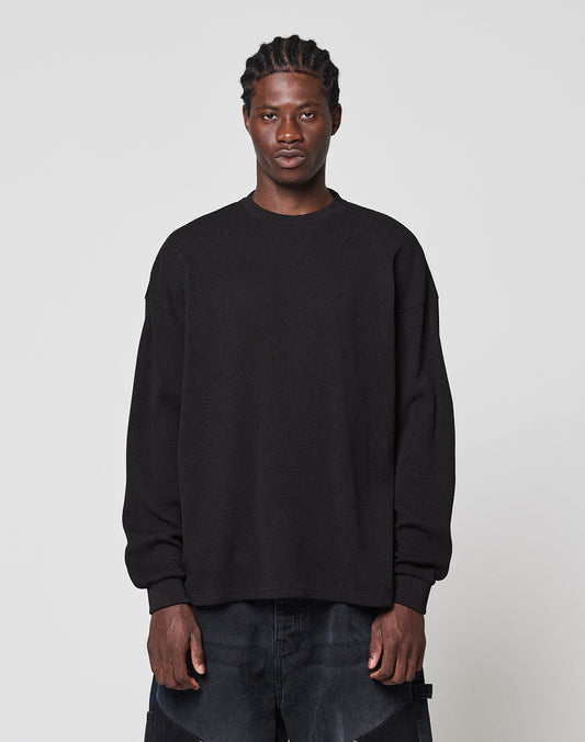 A person with short braided hair and a neutral expression stands against a plain light gray background, wearing the LIVE FAST DIE YOUNG Blank Waffle Longsleeve in black with Ripp-Bündchen and dark pants.