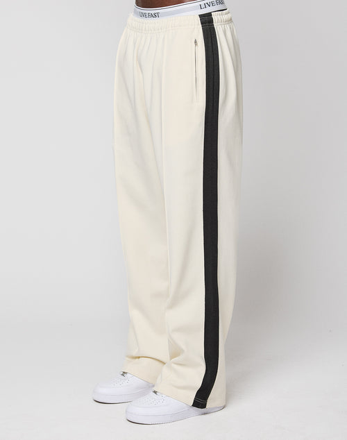 A person wears the LFDY Striped Jogger in cream by LIVE FAST DIE YOUNG, featuring black herringbone stripes, an elastic waistband with "LIVE FAST" text, and white sneakers. The background is plain white.