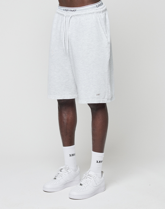 A person wears LIVE FAST DIE YOUNG Basic Shorts in regular fit with elastic waistband and logo print, paired with white LFDY socks and white Nike sneakers, standing against a plain white background.