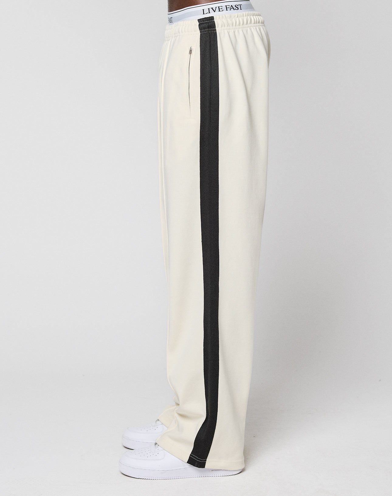 A person in off-white LIVE FAST DIE YOUNG LFDY Striped Jogger with a black side stripe, white sneakers, and a LIVE FAST waistband stands against a plain light background.