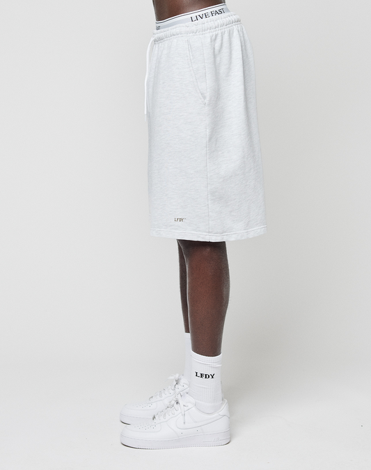 A person stands in profile wearing LIVE FAST DIE YOUNG Basic Shorts in light gray with an elastic waistband and "LIVE FAST" text, plus white socks with LFTY logo print and white sneakers. The shorts have a regular fit. The background is plain white.