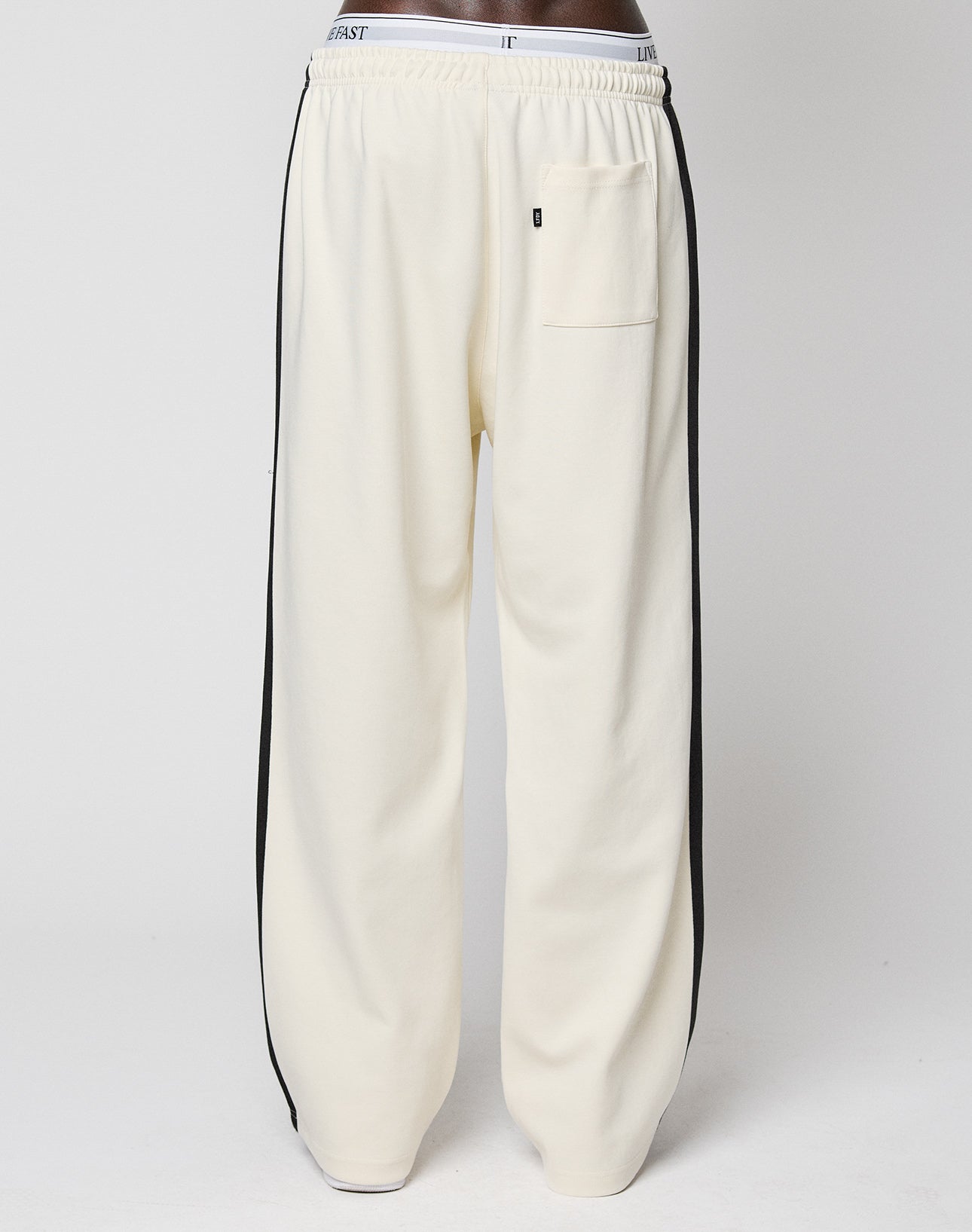 A person wearing the LIVE FAST DIE YOUNG LFDY Striped Jogger in off-white with black herringbone tape side stripes, an elastic waistband, and a small left hip pocket, standing against a plain light background.