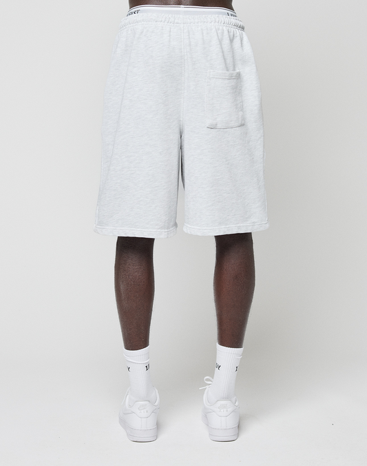 A person faces away, wearing LIVE FAST DIE YOUNG Basic Shorts in light gray with elastic waistband and back pocket, paired with white socks and white sneakers, set against a plain light background.