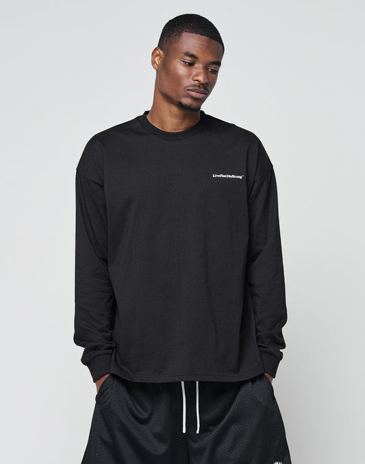 A man stands against a light gray background, looking downward with hands in his pockets, wearing the LIVE FAST DIE YOUNG Basic Longsleeve—an oversized black shirt with white chest logo—and black shorts.