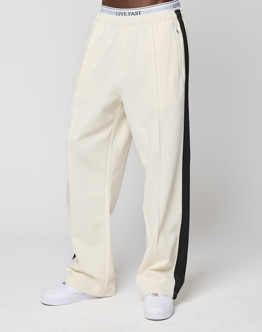 A person wears off-white LFDY Striped Jogger pants by LIVE FAST DIE YOUNG, featuring black side stripes and an elastic waistband with "LIVE FAST" text, paired with white sneakers against a plain white background.
