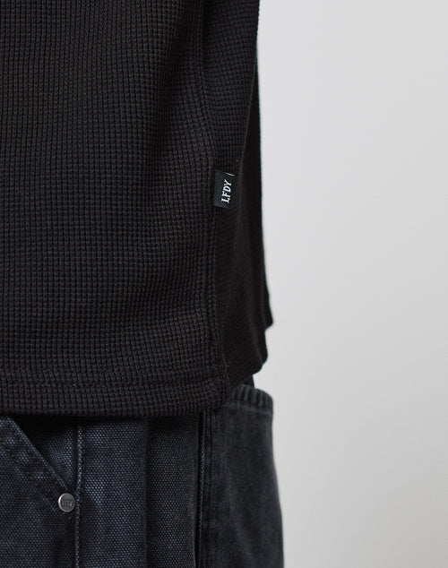 Close-up of a person wearing the LIVE FAST DIE YOUNG Blank Waffle Longsleeve, a black textured shirt with Ripp-Bündchen and a HEMP fabric tag near the hem, paired with dark denim pants against a light plain background.