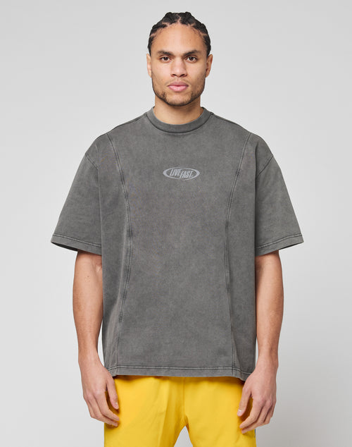 A man with braided hair wears the LIVE FAST DIE YOUNG Live Fast Wave Tee, an oversized, faded gray T-shirt with a small LIVE FAST reflective front print, paired with bright yellow shorts, standing against a light gray background.