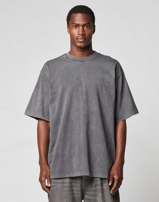A man faces forward against a plain light gray background, wearing the LIVE FAST DIE YOUNG LFDY Hardwear Tee in Oversize Fit, a loose-fitting faded black T-shirt, paired with matching dark jeans.
