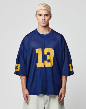 A person with short, bleached blond hair wears the blue LFDY Mesh Jersey by LIVE FAST DIE YOUNG, featuring an oversize fit, yellow number 13 on the front and sleeves, and a V-Ausschnitt, standing against a plain light background.