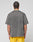 A person with braided hair is seen from the back, wearing the LIVE FAST DIE YOUNG Live Fast Wave Tee in faded gray with an oversize fit, paired with bright yellow shorts, standing against a plain light gray background.