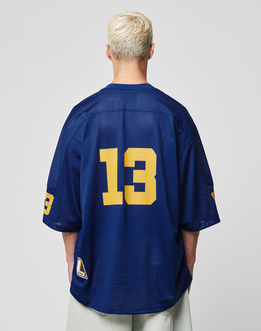 A person with short, bleached blond hair faces away, wearing the LIVE FAST DIE YOUNG LFDY Mesh Jersey in Oversize Fit, navy blue with bold yellow number 13 on back and sleeve. The backdrop is plain light gray.