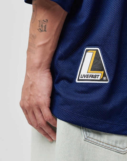 A hand rests beside light-wash jeans, wearing the LIVE FAST DIE YOUNG LFDY Mesh Jersey in navy with an oversize fit and a yellow, black, and white LIVEFAST web patch; initials tattooed on the forearm are also visible.