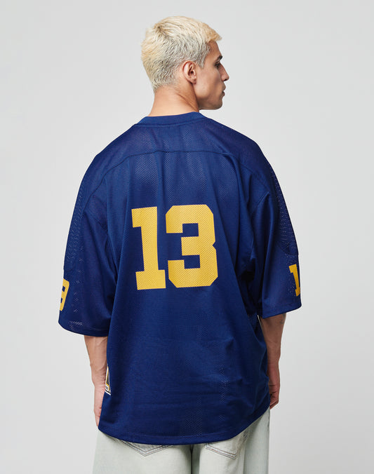 A person with short, bleached blonde hair wears the oversize LFDY Mesh Jersey by LIVE FAST DIE YOUNG. The loose-fit blue polyester jersey features a V-neck and yellow number 13 on the back and sleeves. The person faces away from a plain background.