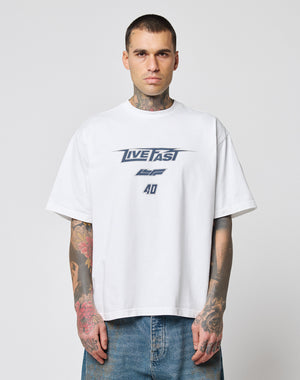 A tattooed man wears the oversize fit LFDY Triple Racing Tee by LIVE FAST DIE YOUNG, a white 100% Baumwolle shirt with LIVE FAST and 40 in blue, paired with blue jeans. He stands against a plain light background, facing the camera.