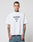A tattooed man wears the oversize fit LFDY Triple Racing Tee by LIVE FAST DIE YOUNG, a white 100% Baumwolle shirt with LIVE FAST and 40 in blue, paired with blue jeans. He stands against a plain light background, facing the camera.