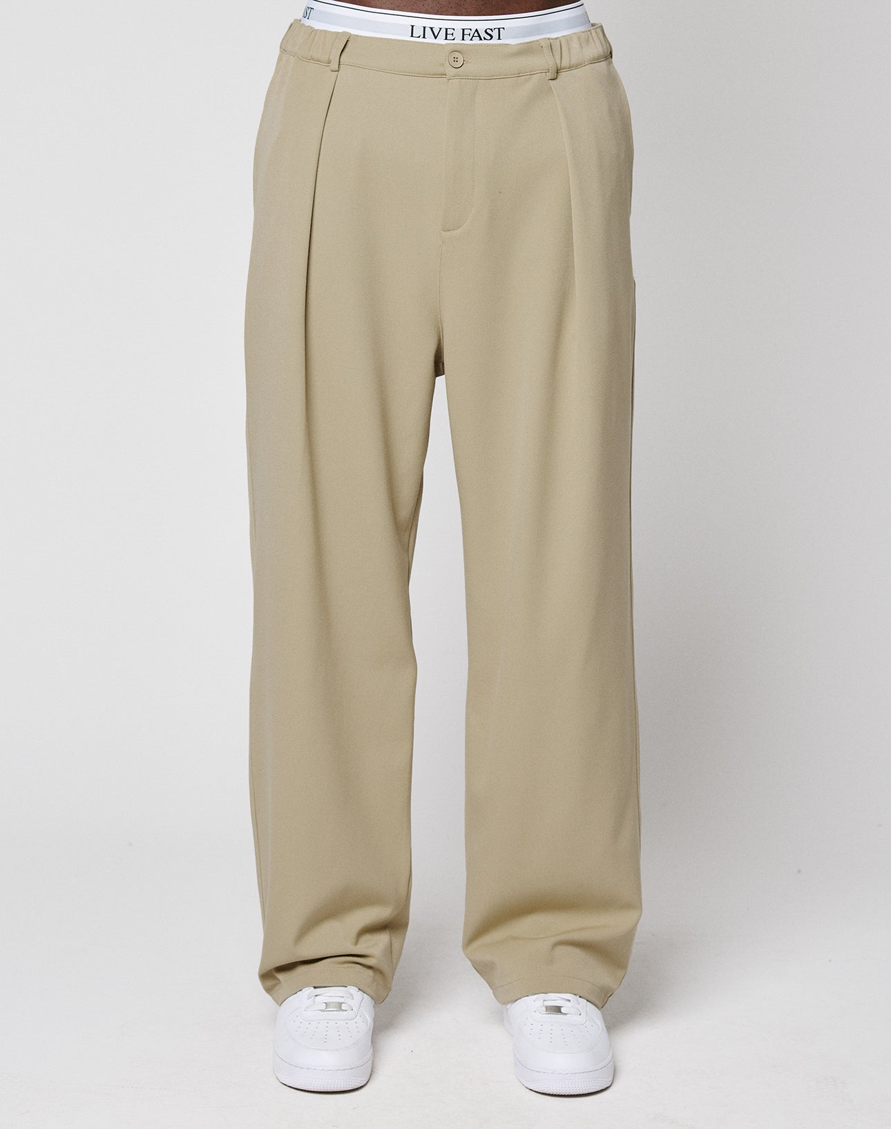 A person models LIVE FAST DIE YOUNG's Wide Casual Pants—beige, pleated, with elastic waistband—paired with white sneakers and visible LIVE FAST-branded underwear, standing against a plain white background.
