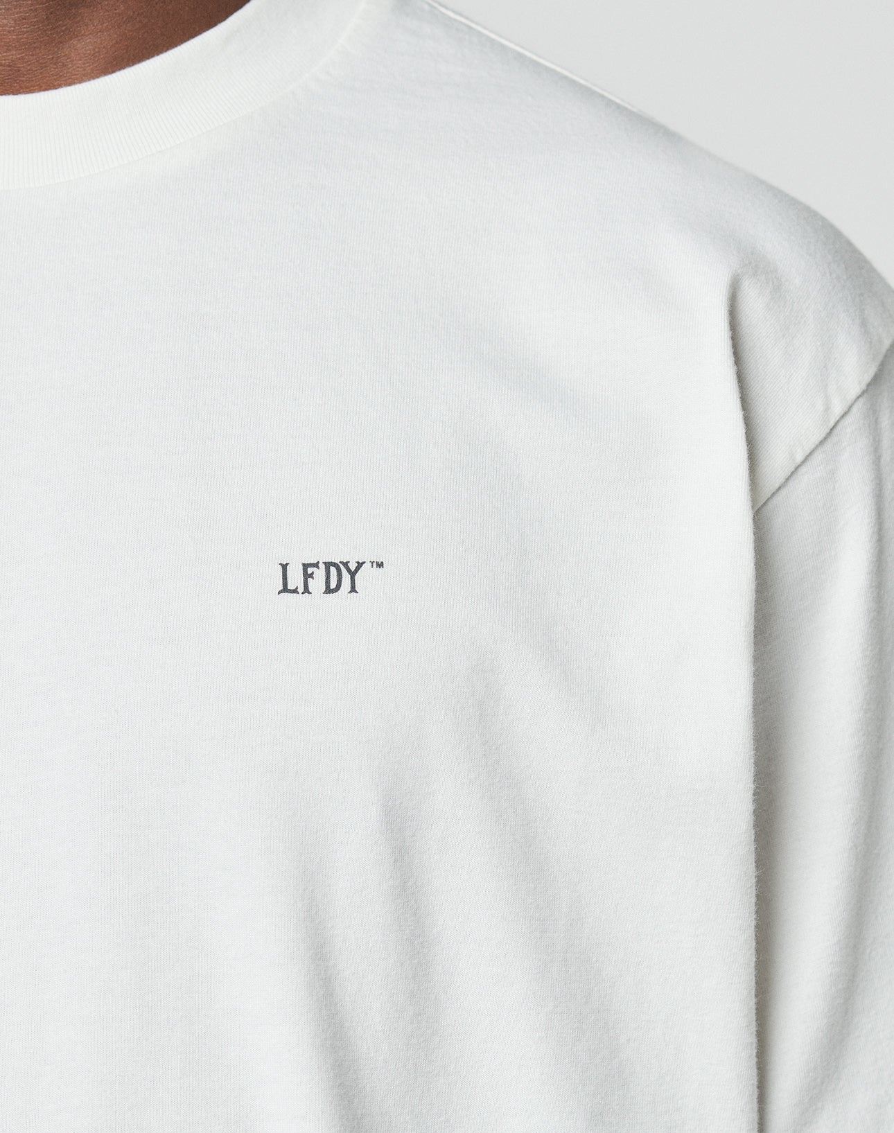 Close-up of a person wearing the LIVE FAST DIE YOUNG LFDY Zizou Tee, an oversized white t-shirt made from 100% cotton, with small black LFDY™ lettering on the left chest. Only the upper shirt, shoulder, and neck are visible.