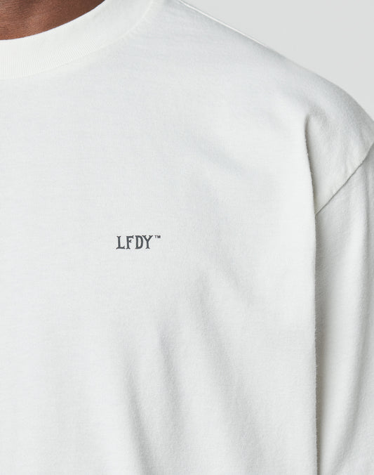 Close-up of a person wearing the LIVE FAST DIE YOUNG LFDY Zizou Tee, an oversized white t-shirt made from 100% cotton, with small black LFDY™ lettering on the left chest. Only the upper shirt, shoulder, and neck are visible.