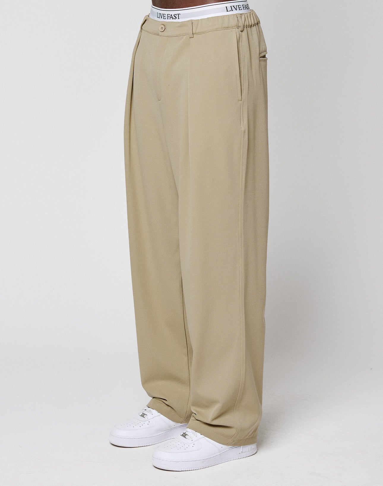 Someone stands against a plain white background wearing LIVE FAST DIE YOUNG Wide Casual Pants in beige with an elastic waistband, paired with white sneakers and a visible white "LIVE FAST" branded waistband.
