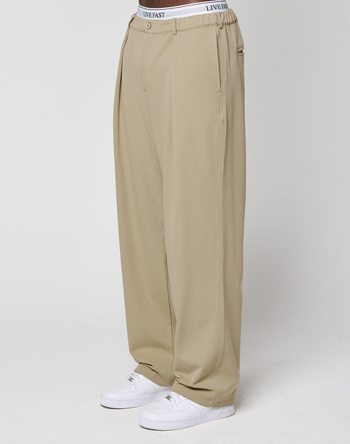 Someone stands against a plain white background wearing LIVE FAST DIE YOUNG Wide Casual Pants in beige with an elastic waistband, paired with white sneakers and a visible white "LIVE FAST" branded waistband.