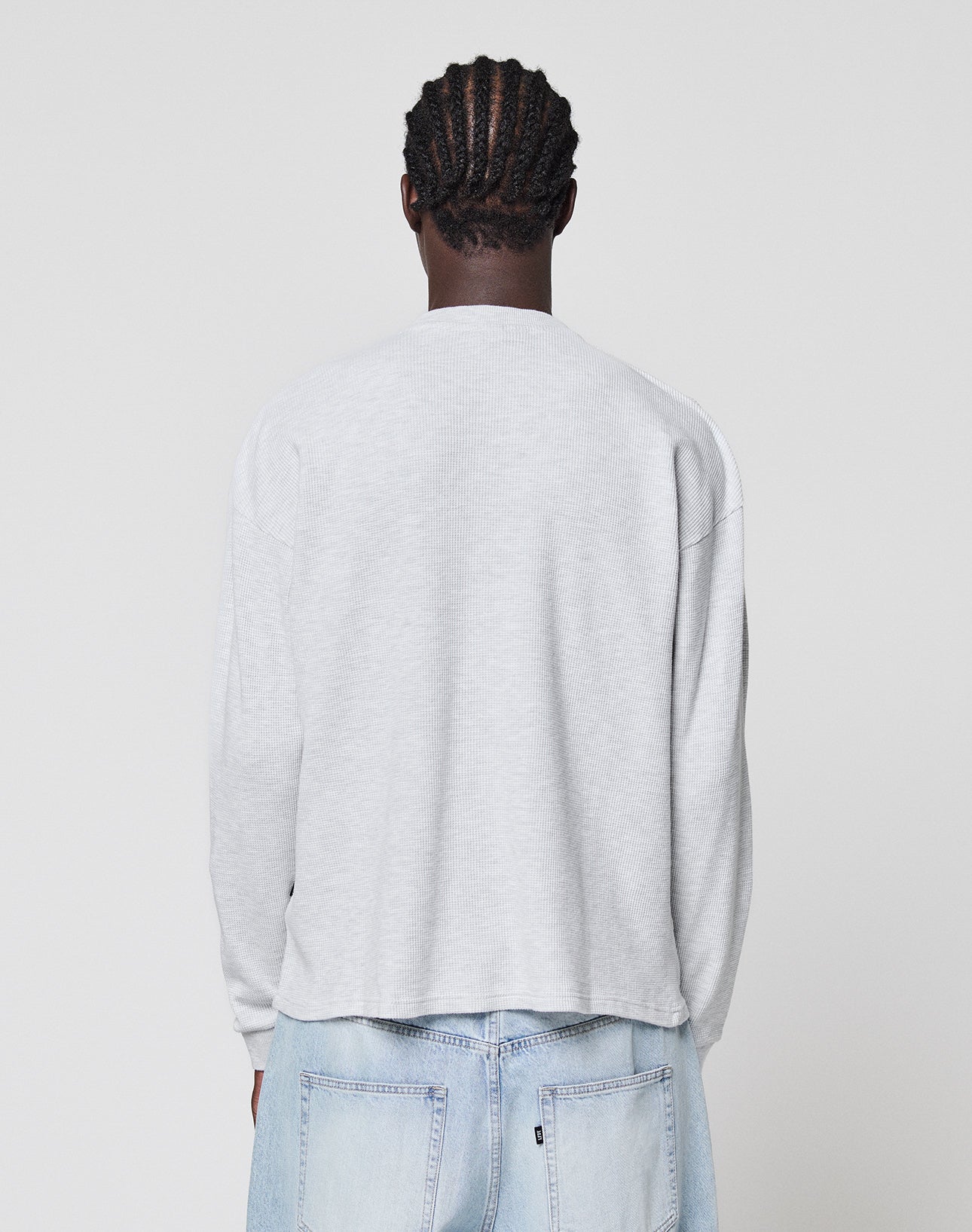 A person with braided hair faces away from the camera, wearing a LIVE FAST DIE YOUNG Blank Waffle Longsleeve in light gray Oversize Fit and light blue jeans against a plain white background.