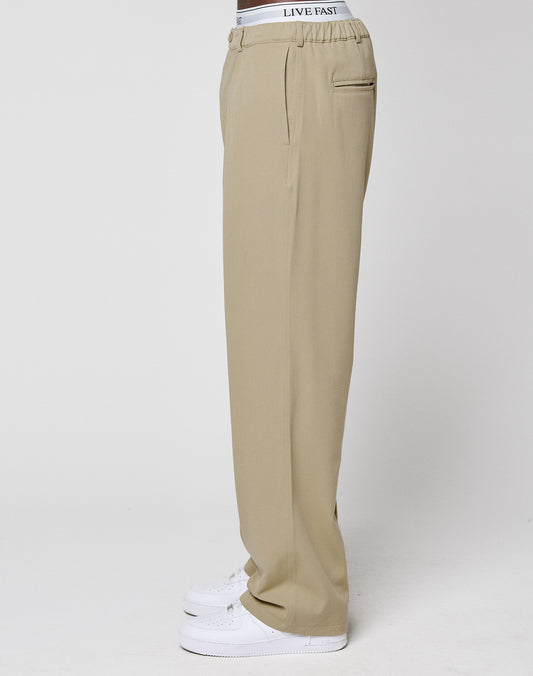 A person stands sideways against a plain white background, wearing LIVE FAST DIE YOUNG Wide Casual Pants in beige with wide legs, a visible white waistband featuring "LIVE FAST" text, and white sneakers.
