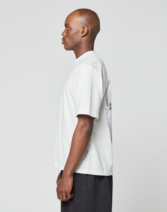 A person stands in profile facing left, wearing the LIVE FAST DIE YOUNG LFDY Zizou Tee in white (100% Baumwolle, oversize fit, front and backprint) with dark pants against a plain light gray background; the backprint is partially visible.