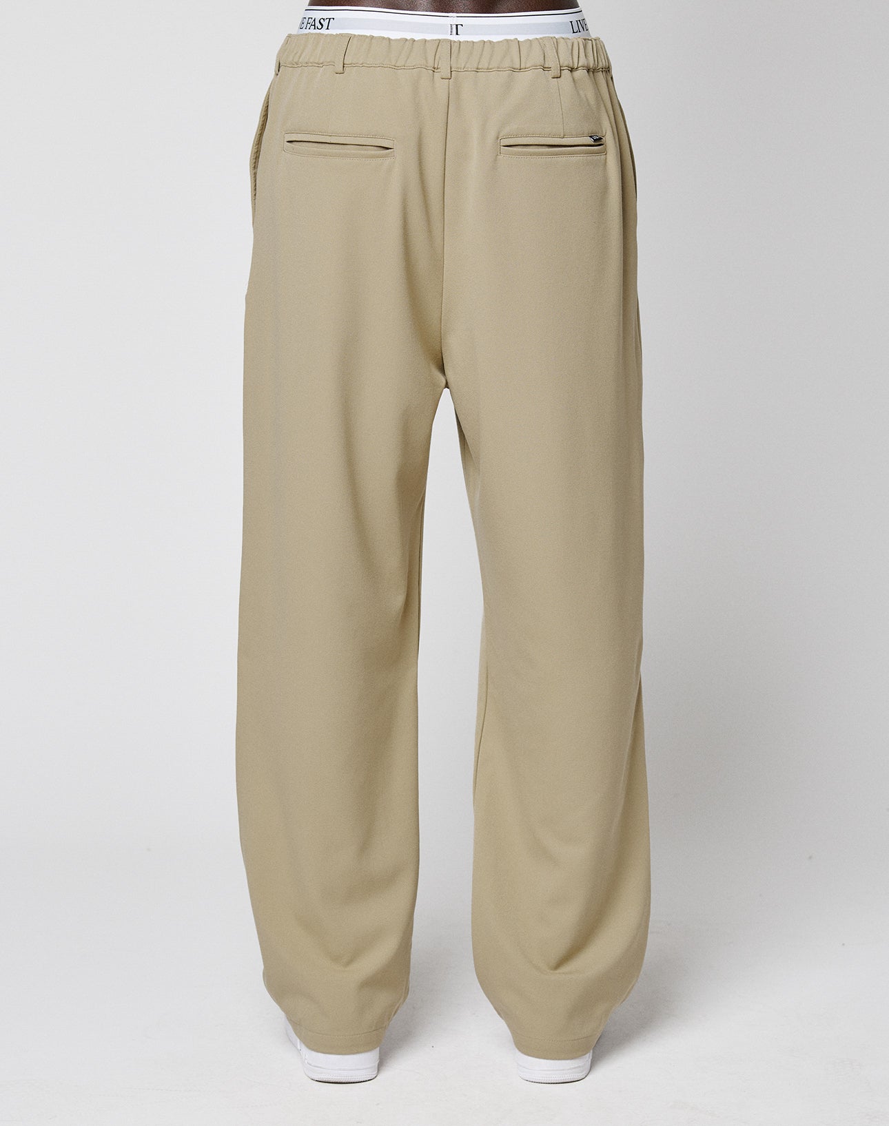A person stands against a plain light background wearing LIVE FAST DIE YOUNG Wide Casual Pants in beige, with an elastic waistband, wide legs, and two back welt pockets.