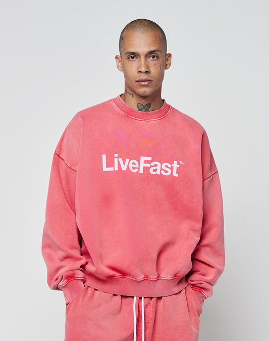 A person with a shaved head wears the LIVE FAST DIE YOUNG Basic LiveFast Sweaterāan oversized pink sweatshirt featuring a bold white LiveFast front printāwhile standing against a plain light background.