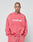 A person with a shaved head wears the LIVE FAST DIE YOUNG Basic LiveFast Sweater—an oversized pink sweatshirt featuring a bold white LiveFast front print—while standing against a plain light background.