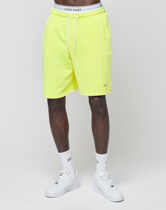 A person wears LIVE FAST DIE YOUNG Basic Shorts in neon yellow with "LFT" logo print and a white waistband reading "LIVE FAST", paired with white socks and sneakers, standing against a plain light background.