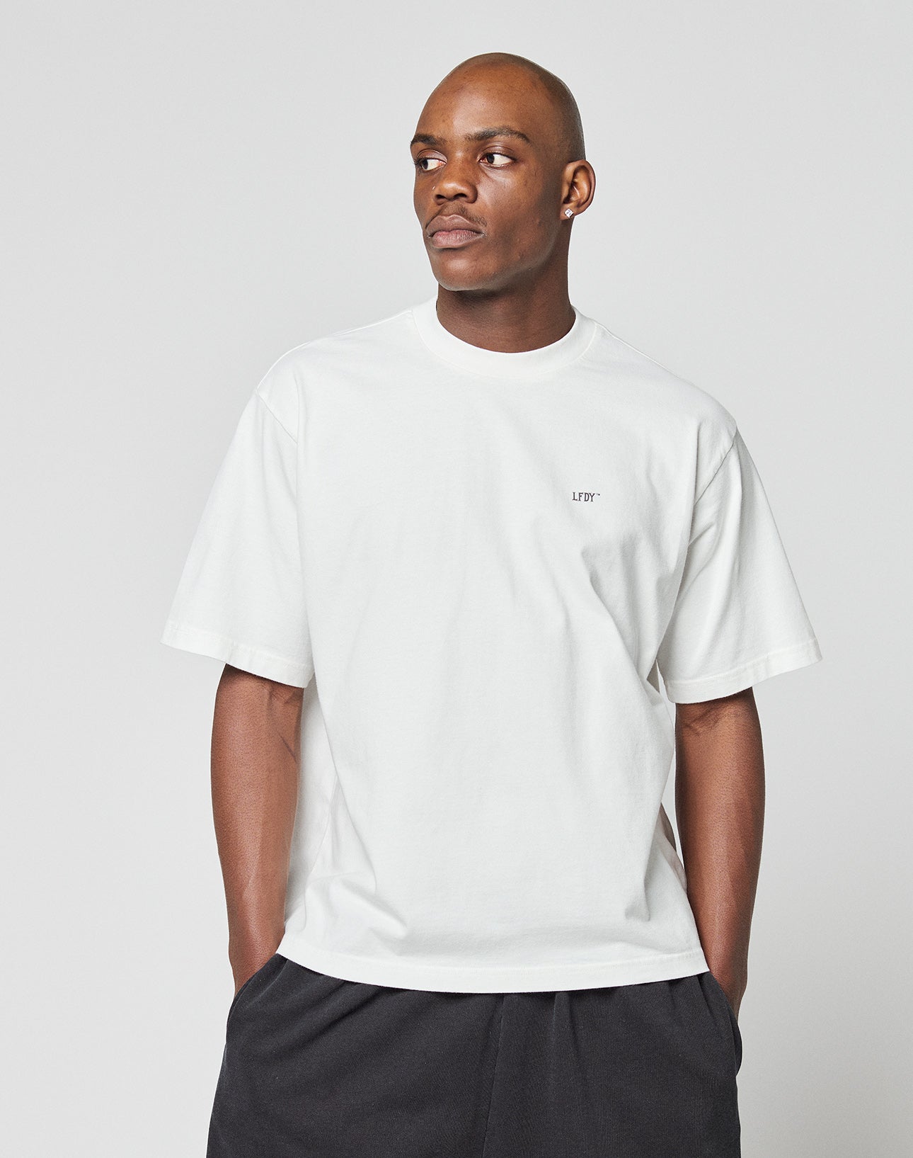 A person wears the LFDY Zizou Tee by LIVE FAST DIE YOUNG—an oversize white 100% Baumwolle T-shirt with small LFDY chest text—and black pants, standing against a plain light gray background looking to the side.