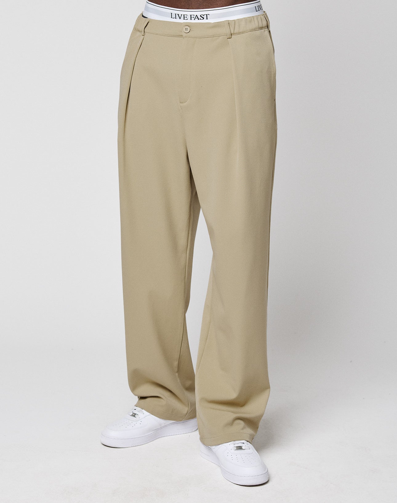 A model wears LIVE FAST DIE YOUNG Wide Casual Pants in beige (wide fit, elastic waistband), white sneakers, and visible white LIVE FAST underwear, standing against a plain white background.