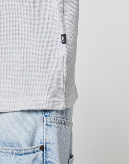 Close-up of the LIVE FAST DIE YOUNG Blank Waffle Longsleeve in light gray with a textured, oversize fit, worn over light blue denim jeans. The shirt features a small black flag label. Background is plain and light-colored.