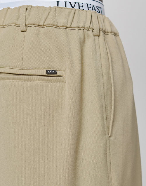 Close-up of beige Wide Casual Pants by LIVE FAST DIE YOUNG, featuring a back pocket, side seam, and a small metallic tag. The waistband of white underwear branded LIVE FAST is visible above the pants.