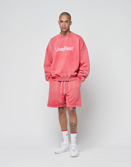 A person with short hair and visible neck and leg tattoos wears a pink Basic LiveFast Sweater by LIVE FAST DIE YOUNG, matching pink shorts, white crew socks, and white sneakers, standing against a plain background.
